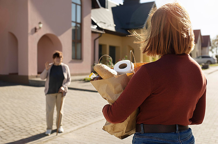 Jüngere Person mit Einkaufstasche, Brot und Toilettenpapier vor älterer Person mit Stock